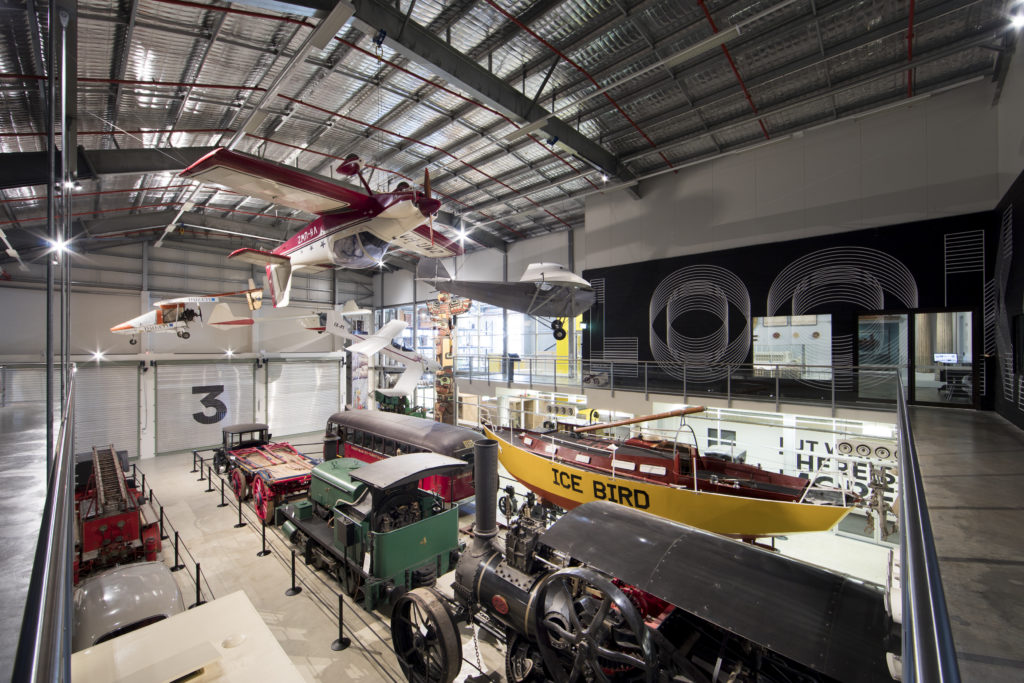View overlooking a gallery space at the Museums Discovery Centre in Castle Hill. Hanging from the ceiling are several small planes, including one which is upside down. On the level below are other objects from the Museum's transport collection, including a boat, tram carriage, steam train and fire engine.